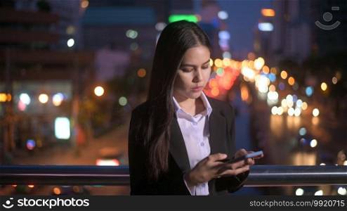 A young beautiful business woman is using smart phone in city over traffic lights at evening. business and technology concept. 