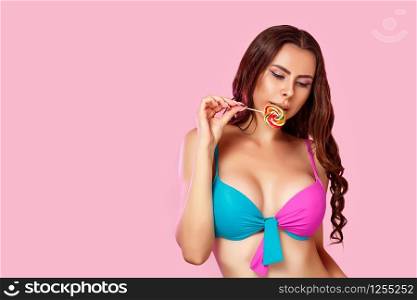 a young beautiful girl in a bright swimsuit licks a colorful bright Lollipop photographed in close-up. on a pink isolated background. with copyspace