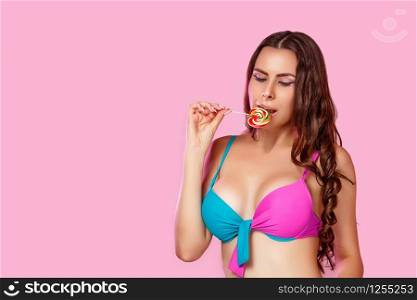 a young beautiful girl in a bright swimsuit licks a colorful bright Lollipop photographed in close-up. on a pink isolated background. with copyspace