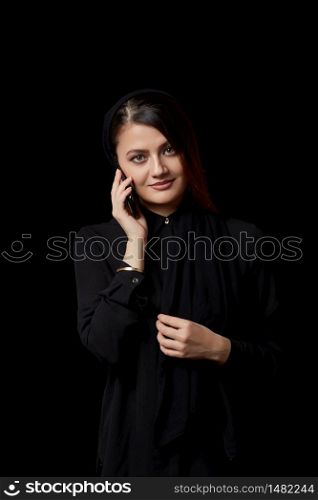 a young beautiful Muslim woman in black clothes uses a mobile phone.smiling, texting, talking. on a black isolated background.