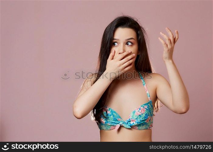a young beautiful slender Asian girl in a swimsuit covered her mouth with her hand in surprise, in admiration, in delight . on a pink isolated background.
