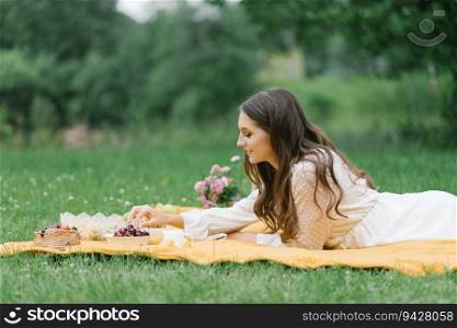 A young beautiful woman in a white dress sits on a blanket on a summer picnic on the green grass. Happy spending time in nature in the summer