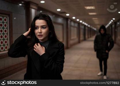 a young beautiful woman walks through an underground passage at night, followed by a man in dark clothes with a hood on his head