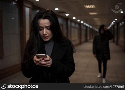a young beautiful woman walks through an underground passage at night, followed by a man in dark clothing with a hood on his head. a woman tries to dial the number of the rescue service on the phone