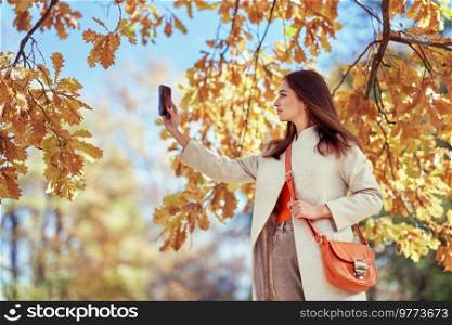 A young beautiful woman walks through the autumn park. A girl with a smartphone takes photos in an autumn forest