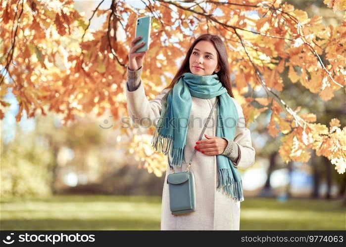 A young beautiful woman walks through the autumn park. A girl with a smartphone takes photos in an autumn forest