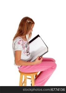 A young blond girl sitting in pink pants, holding her folderand writing in her book, isolated for white background.
