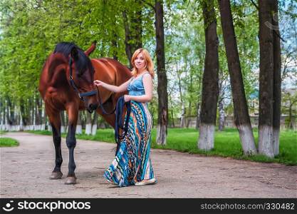 A young blond woman in a long blue dress holds the reins of her horse and smiling at the camera against the backdrop of beautiful green trees.. Young blond with a beautiful horse