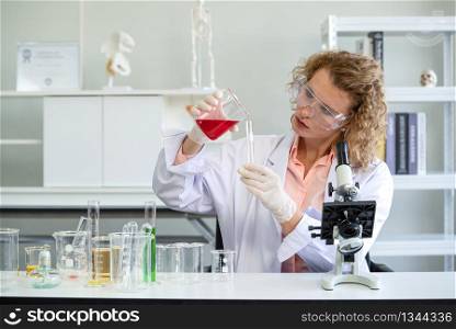 A young blonde scientist is pouring red chemicals into a test tube. Working atmosphere in chemical laboratory. Test tubes and beakers filled with chemicals on the table.