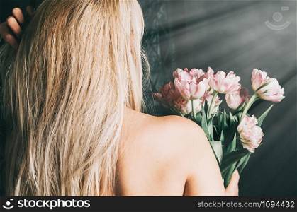 A young blonde woman holding a bouquet of fresh pink flowers by the window, view from her back, bare shoulders. Soft focus image. Romantic, love, happiness concept