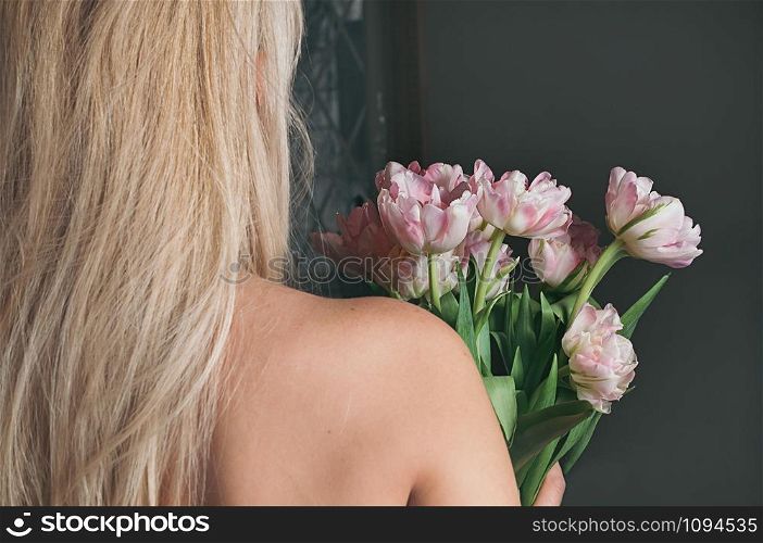 A young blonde woman holding a bouquet of fresh pink flowers by the window, view from her back, bare shoulders