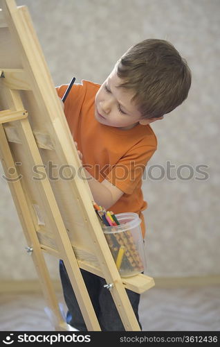 A young boy drawing onto a canvas with a pot of coloured pencils stood on the ledge of the canvas