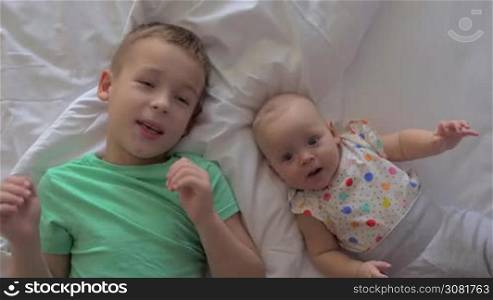 A young boy in a green T-shirt is lying on a back on a bed next to his baby sister. He is moving his hand trying to copy baby&acute;s cute movements. A six months old girl is wearing a polka dot shirt and grey pants