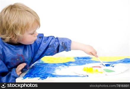 A young boy painting with blue poster paint, reaching for a palette with his brush