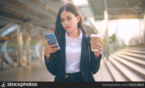 A young business woman wearing black suit is using smart phone , holding a cup of coffee in the city, Business Lifestyle Concept