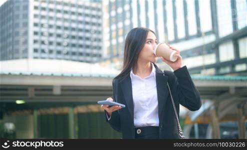A young business woman wearing black suit is using smart phone , holding a cup of coffee in the city, Business Lifestyle Concept