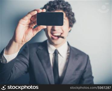 A young businessman is smoking a cigar and is taking a selfie using his smartphone