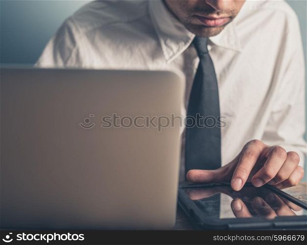 A young businessman is using a tablet while typing on a laptop computer