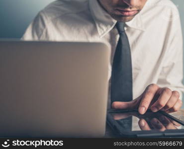 A young businessman is using a tablet while typing on a laptop computer