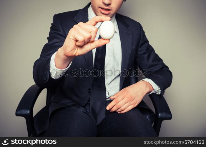 A young businessman sitting in an office chair is holding a boiled egg