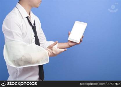 A young businessman with an injured arm in a sling using a tablet over blue background in studio, insurance and healthcare concept. Young businessman with an injured arm in a sling using a tablet over blue background in studio, insurance and healthcare concept
