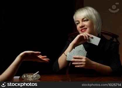 a young Caucasian blonde in black clothes is sitting at a table and playing cards with another woman for undressing and Smoking cigarettes.
