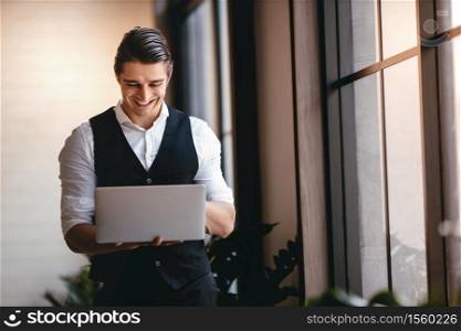 a Young Caucasian Businessman Working on Computer Laptop in the Modern Workplace. Smiling Businessman Standing by the Window