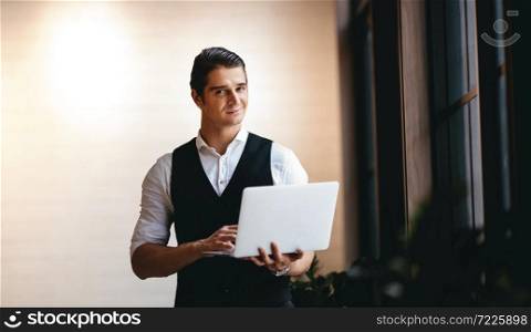 a Young Caucasian Businessman Working on Computer Laptop in the Modern Workplace. Smiling Businessman Standing by the Window and Looking into the Camera