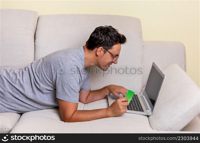A young Caucasian male lying on a couch using a laptop computer and credit card shopping online