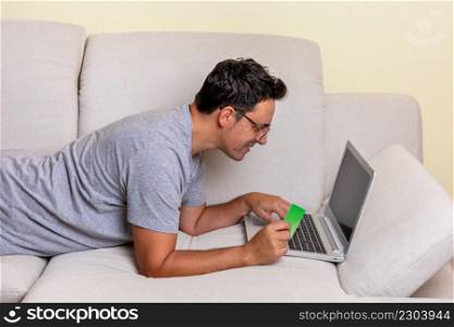 A young Caucasian male lying on a couch using a laptop computer and credit card shopping online