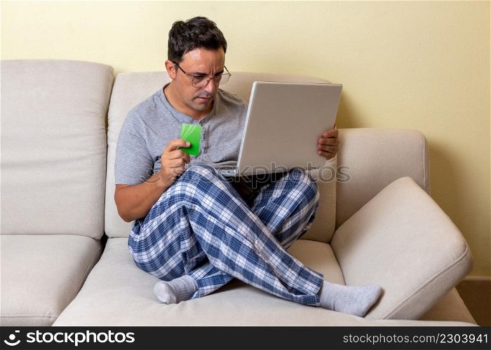 A young Caucasian male sitting on a couch using a laptop computer and credit card shopping online