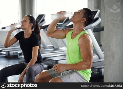 A young Caucasian muscular man drinking water from bottle at gym and blur woman on background