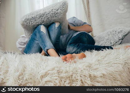 A young couple in blue jeans and shirts having fun on a white bed with grey pillows