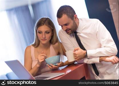 A young couple is preparing for the job and using a laptop. The man drinks coffee while the woman eats breakfast at luxury home together, looking at screen, smiling.