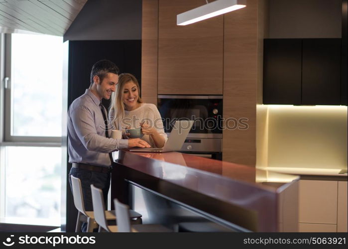 A young couple is preparing for the job and using a laptop. The man drinks coffee while the woman eats breakfast at luxury home together, looking at screen, smiling.