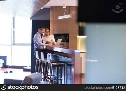 A young couple is preparing for the job and using a laptop. The man drinks coffee while the woman eats breakfast at luxury home together, looking at screen, smiling.