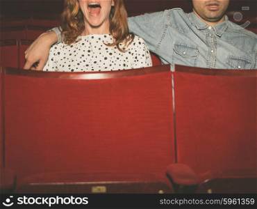 A young couple is watching a film in a movie theater, the woman is scared but her partner is looking relaxed
