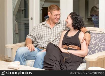 A young couple sit togehter on a bench out on the patio, man has arm around women,s neck, they look at each other while smiling and laughing happily.