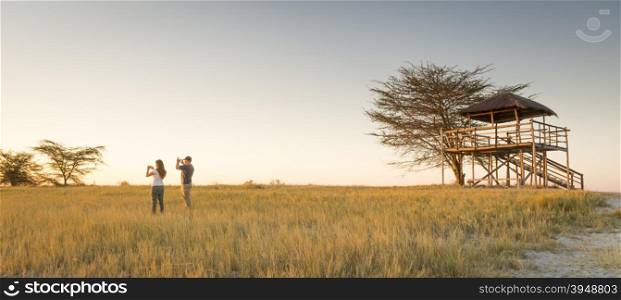 A young couple stand in long grass and take photos of sunset while on safari in the Makgadikgadi Pans, Botswana, Africa