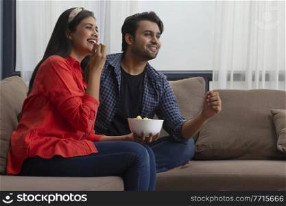 A young couple watching something while eating popcorn in their room.