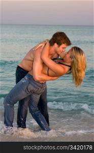 A young couple wearing bluejeans embrace in the surf at sunset.