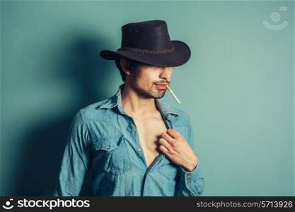 A young cowboy is standing against a blue wall and is smoking a cigarette