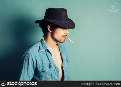 A young cowboy is standing by a blue wall and is smoking a cigarette