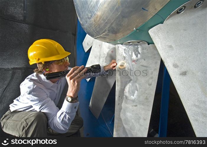 A young engineer inspecting the blades of an industrial windtunnel wearing a hard top, earplugs and protective goggles