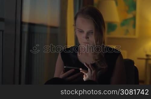 A young fair-haired woman is sitting in some cozy interior in warm shadowed light. She is looking at a smartphone that she is holding in hands with a slight smile
