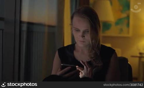 A young fair-haired woman is sitting in some cozy interior in warm shadowed light. She is looking at a smartphone that she is holding in hands with a slight smile