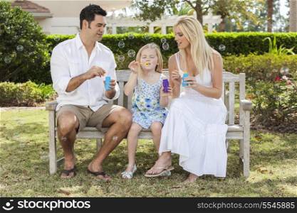 A young family mother &amp; father parents with girl child blowing bubbles having fun together sitting on a bench in a sunny park or garden.