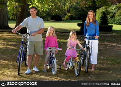 A young family with mother father and two blond daughters riding their bikes in a sun bathed green park