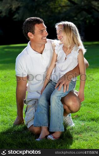 A young father with his blond daughter on his knee having fun and smiling at each other in a sun bathed green park