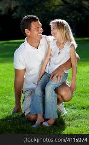 A young father with his blond daughter on his knee having fun and smiling at each other in a sun bathed green park